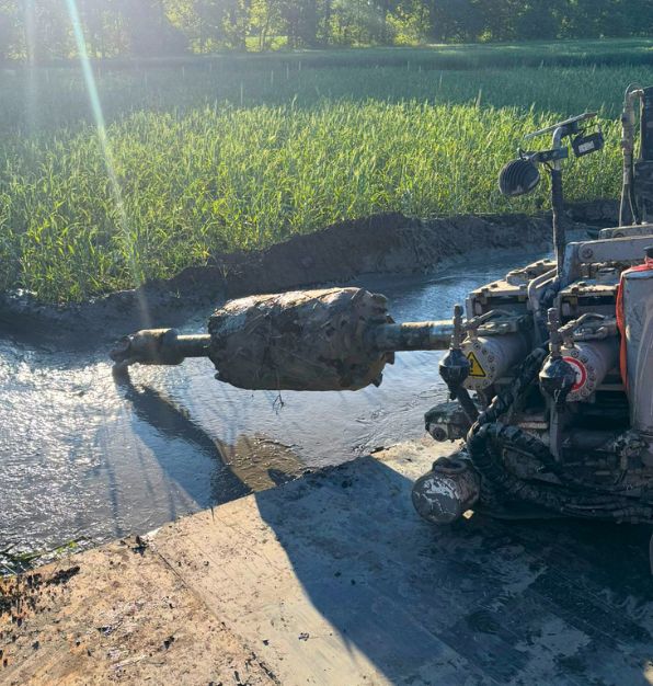 Mud-covered barrel reamer attached to drilling equipment at a muddy worksite, with a grassy field and trees in the background.